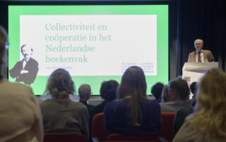 Foto van voorzitter Wouter van Gils tijdens de opening van het symposium. De voorzitter staat achter een katheder en naast hem is een scherm met daarop een dia met de titel van het symposium.